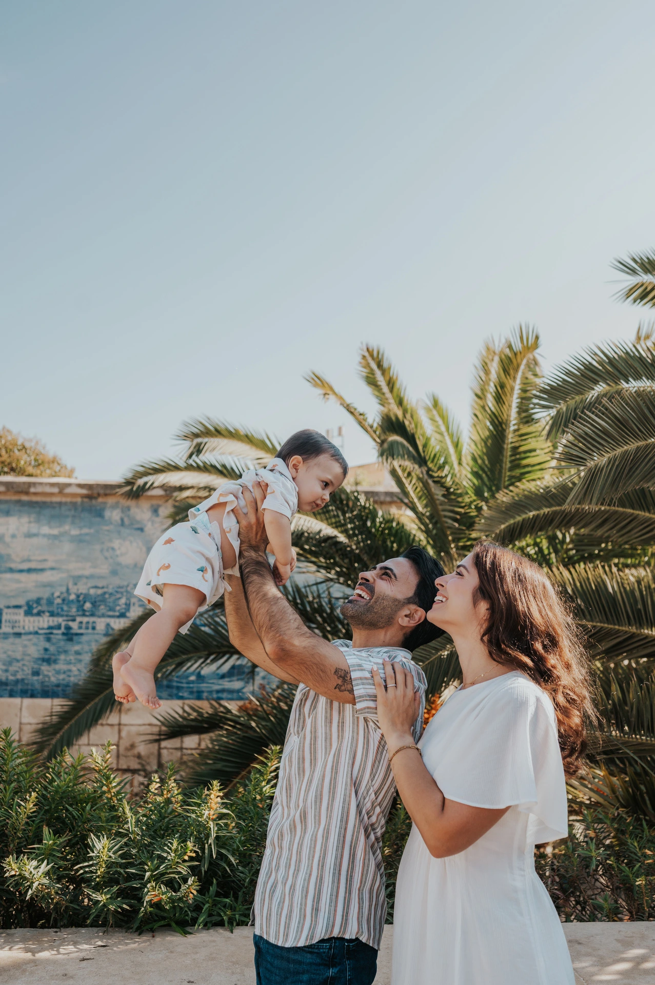 The parents lifting their smiling child between them beside palms and blue tiles during their family photoshoot in Lisbon.
