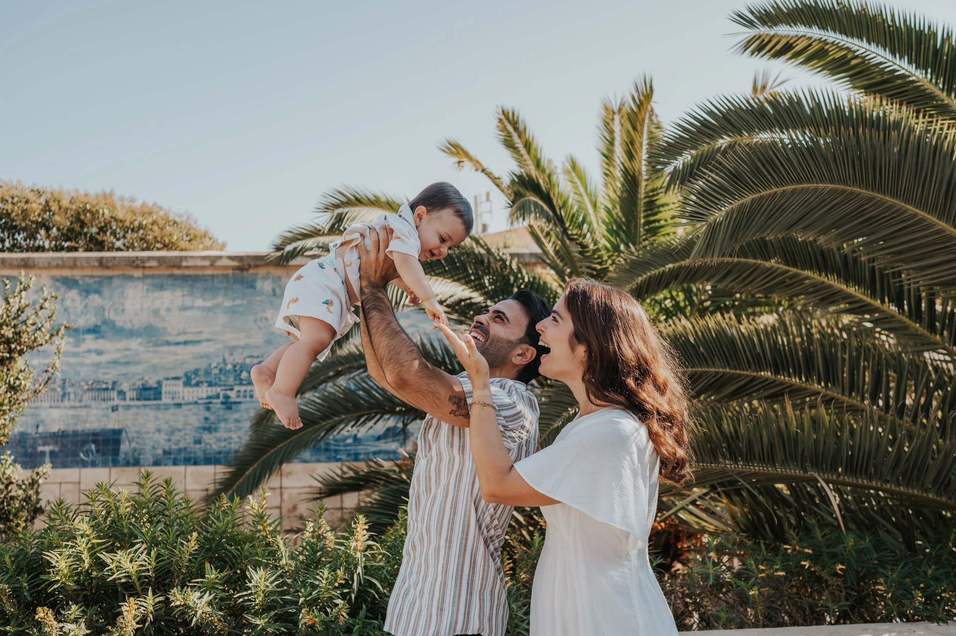 A playful lift beside the palms | Lisbon family photoshoot The parents lifting their smiling child between them beside palms and blue tiles during their family photoshoot in Lisbon.