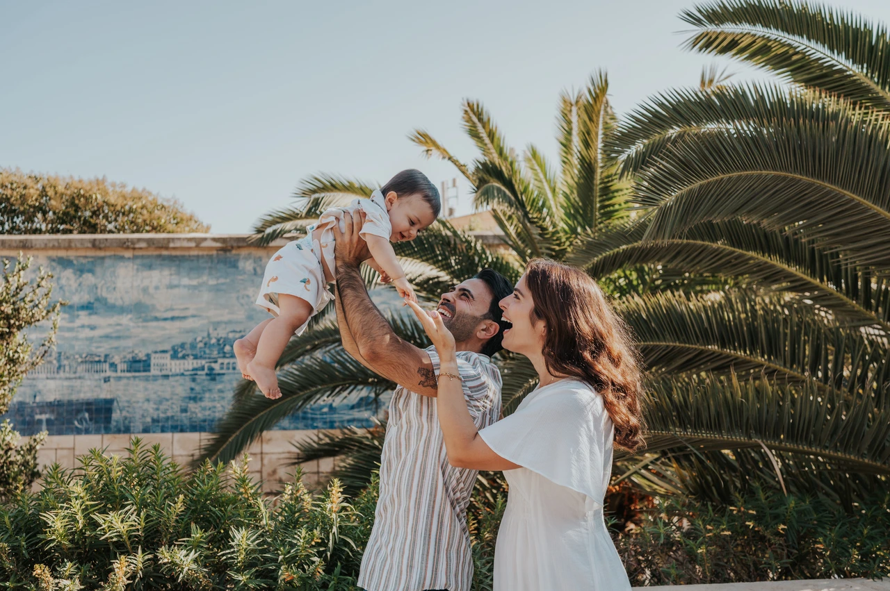 A playful lift beside the palms | Lisbon family photoshoot The parents lifting their smiling child between them beside palms and blue tiles during their family photoshoot in Lisbon.