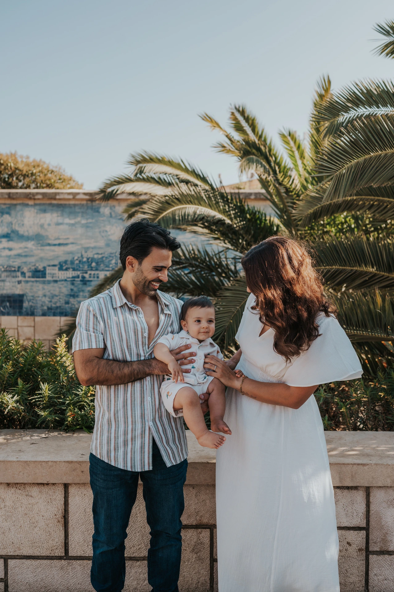 The parents holding their child while standing beside the palm-lined viewpoint during their family photoshoot in Lisbon.