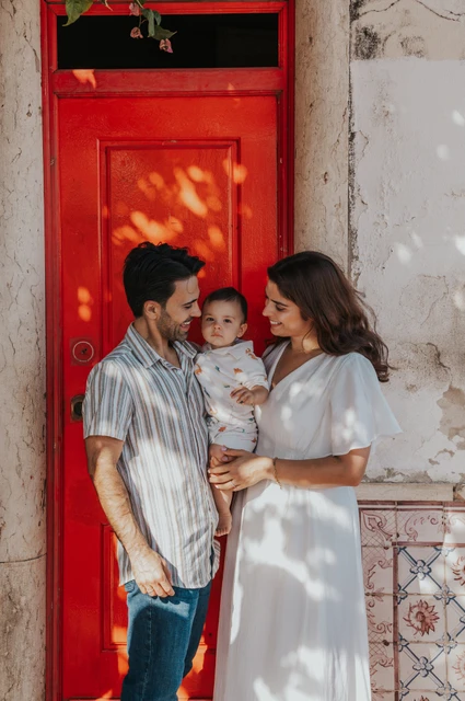 A close family portrait at the red door | Lisbon family photoshoot The parents holding their child close together beside a red doorway during their family photoshoot in Lisbon.