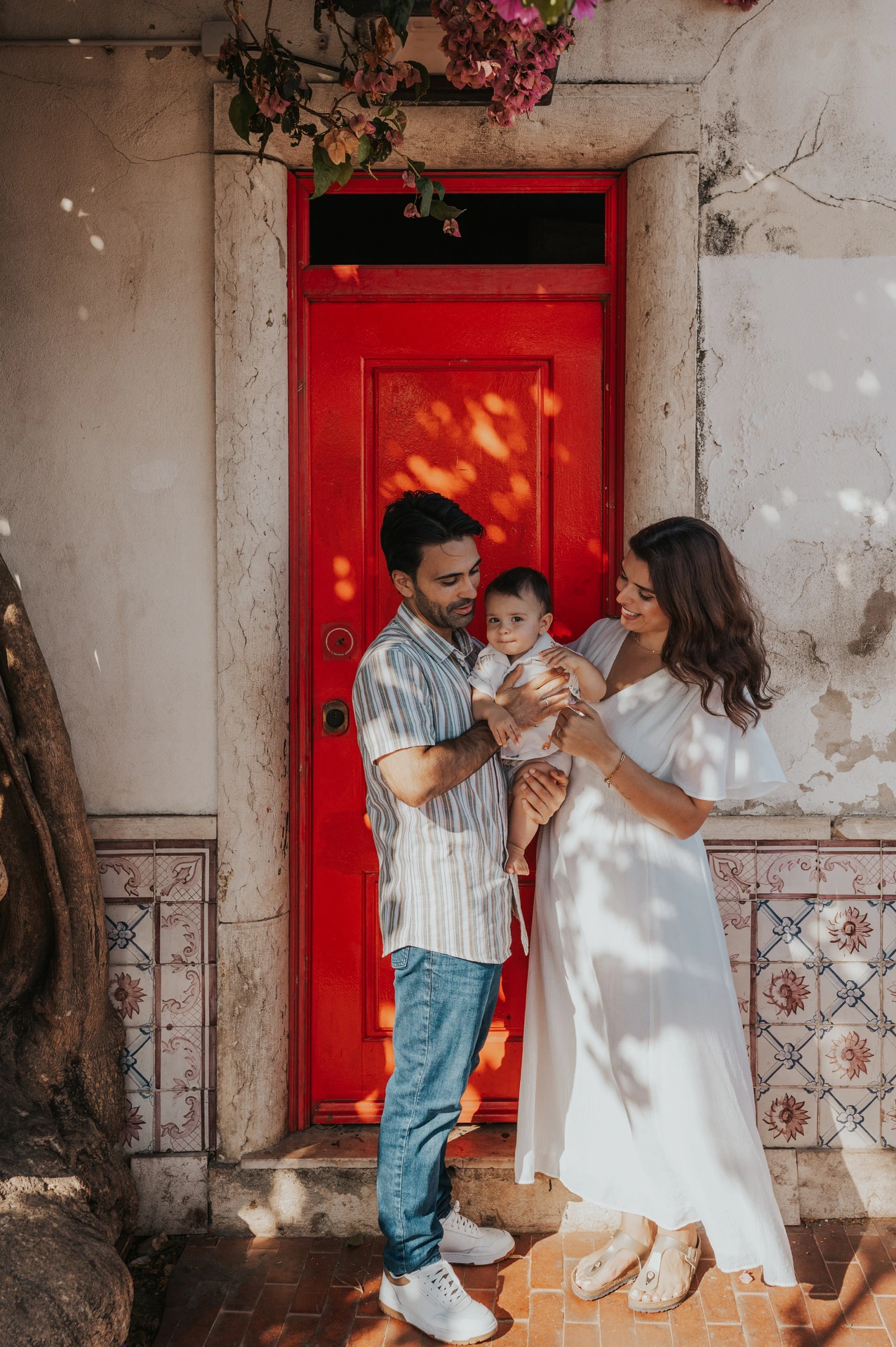 The parents holding their child close together beside a red doorway during their family photoshoot in Lisbon.