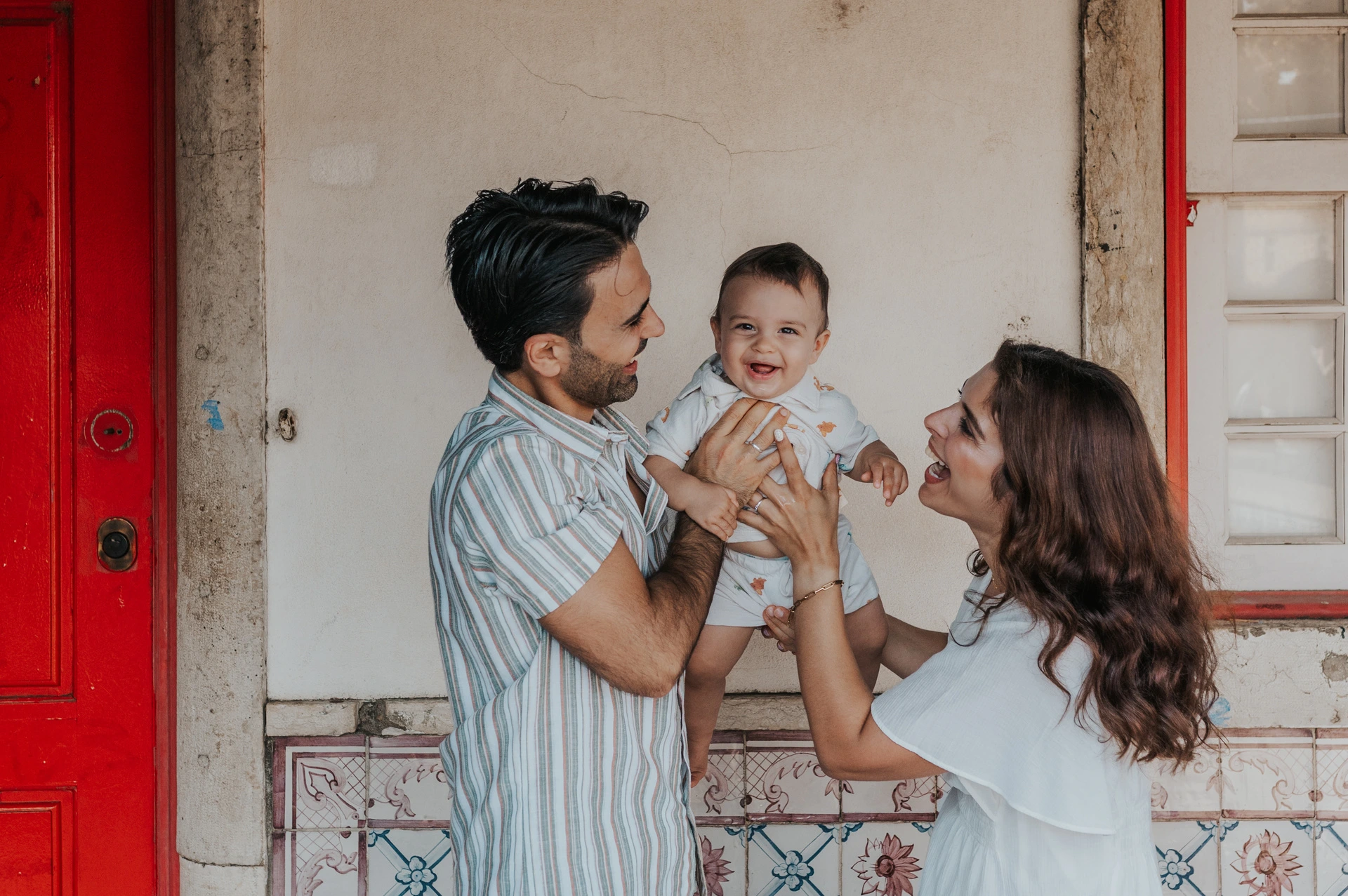 The parents holding their child close together beside a red doorway during their family photoshoot in Lisbon.