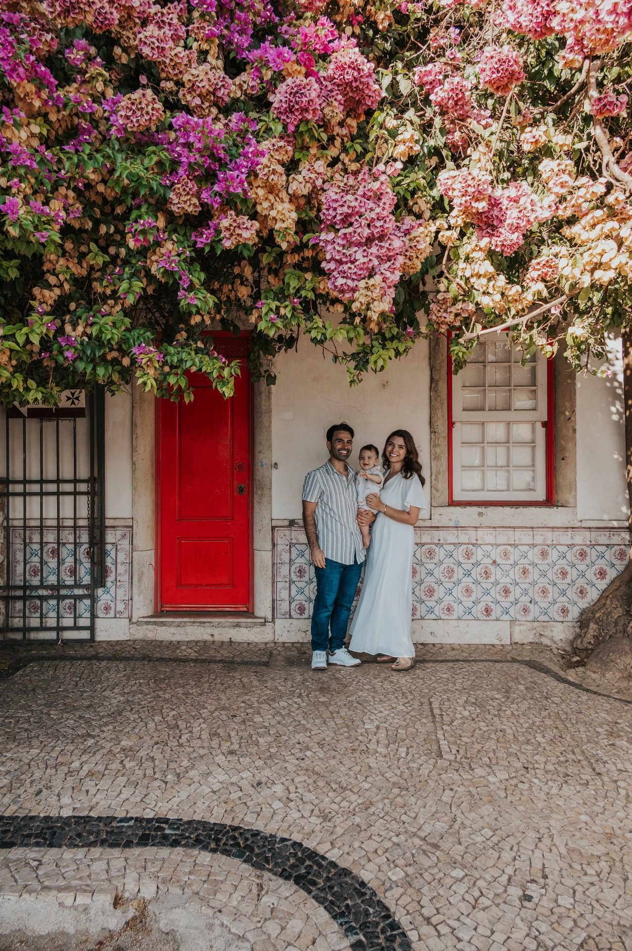 The family standing together at a red doorway beneath flowering branches during their family photoshoot in Lisbon.