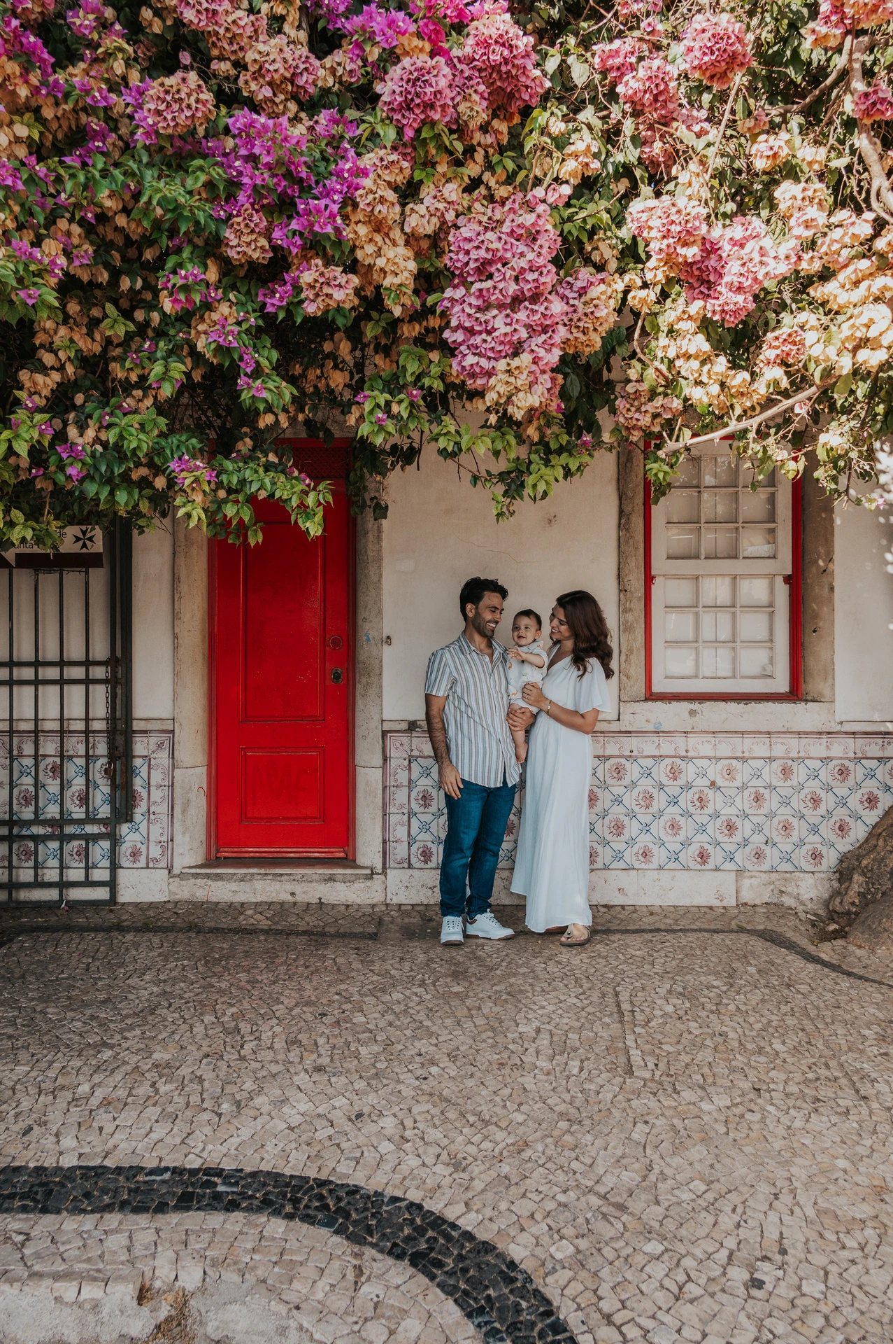 The family standing together at a red doorway beneath flowering branches during their family photoshoot in Lisbon.