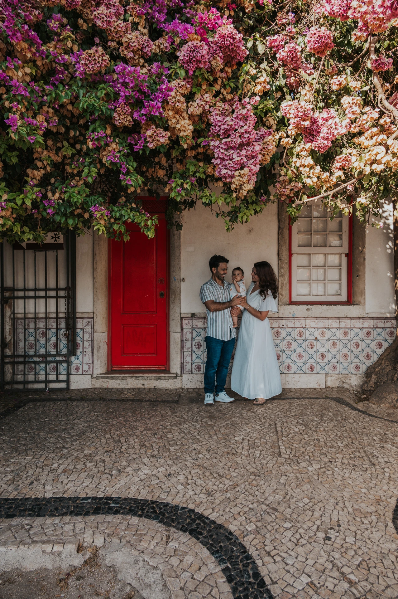The family standing together at a red doorway beneath flowering branches during their family photoshoot in Lisbon.