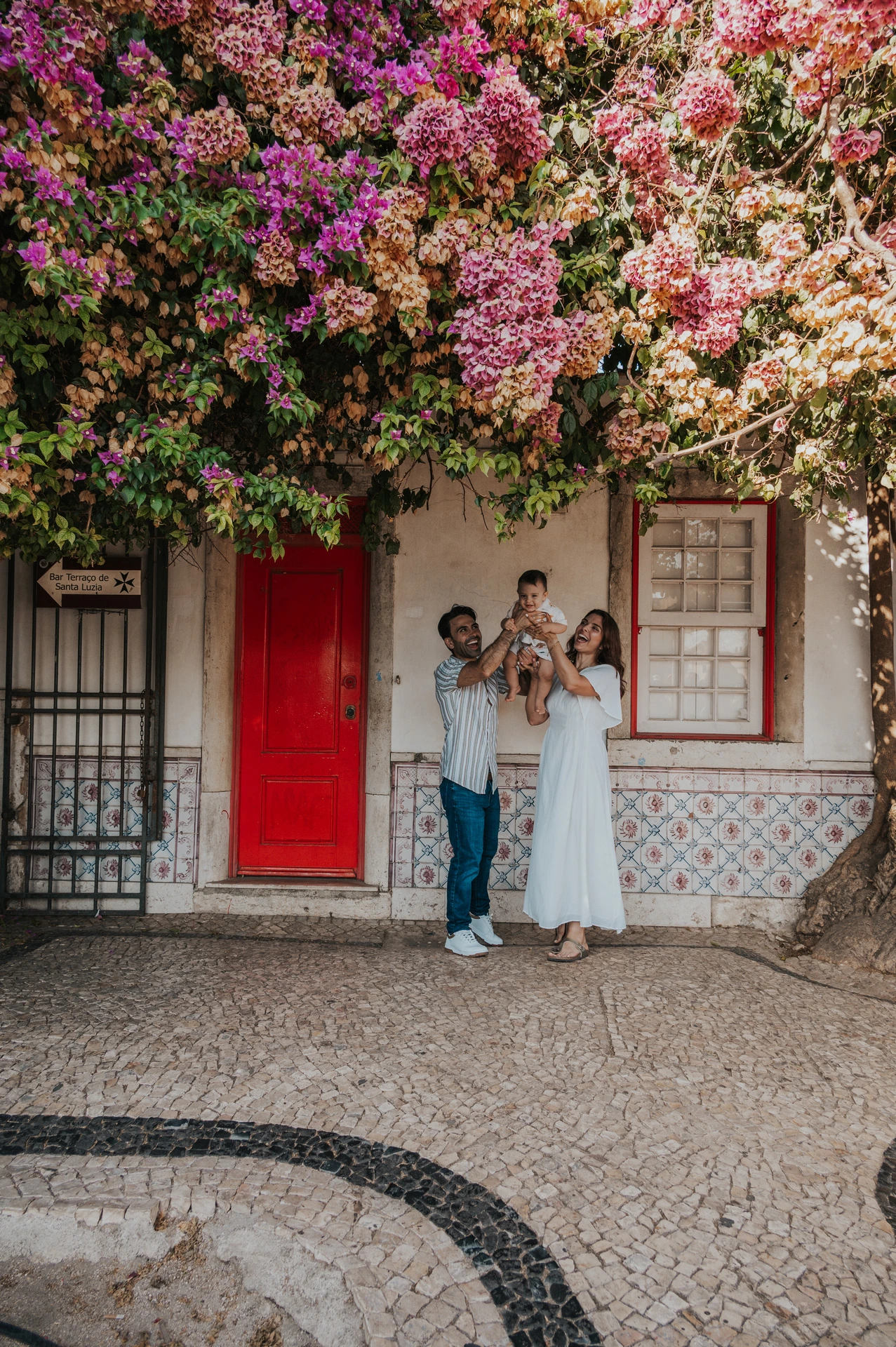The family standing together at a red doorway beneath flowering branches during their family photoshoot in Lisbon.