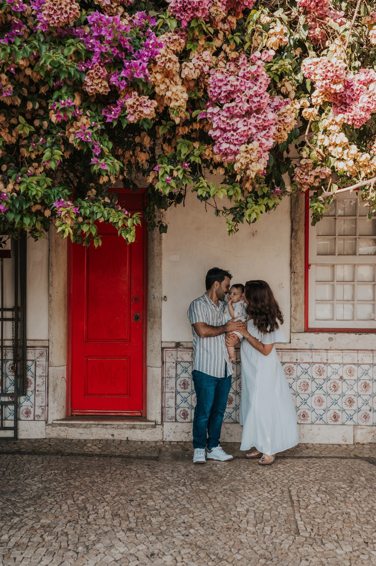 The family standing together at a red doorway beneath flowering branches during their family photoshoot in Lisbon.