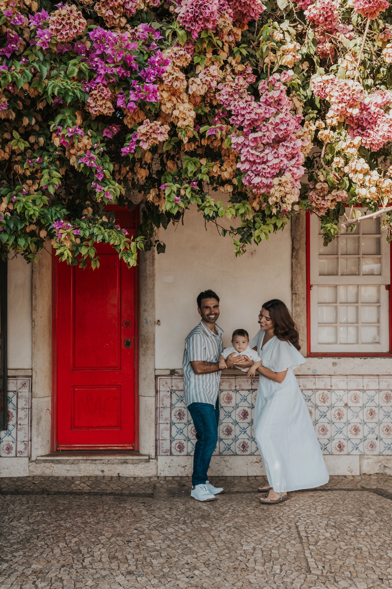 The family standing together at a red doorway beneath flowering branches during their family photoshoot in Lisbon.