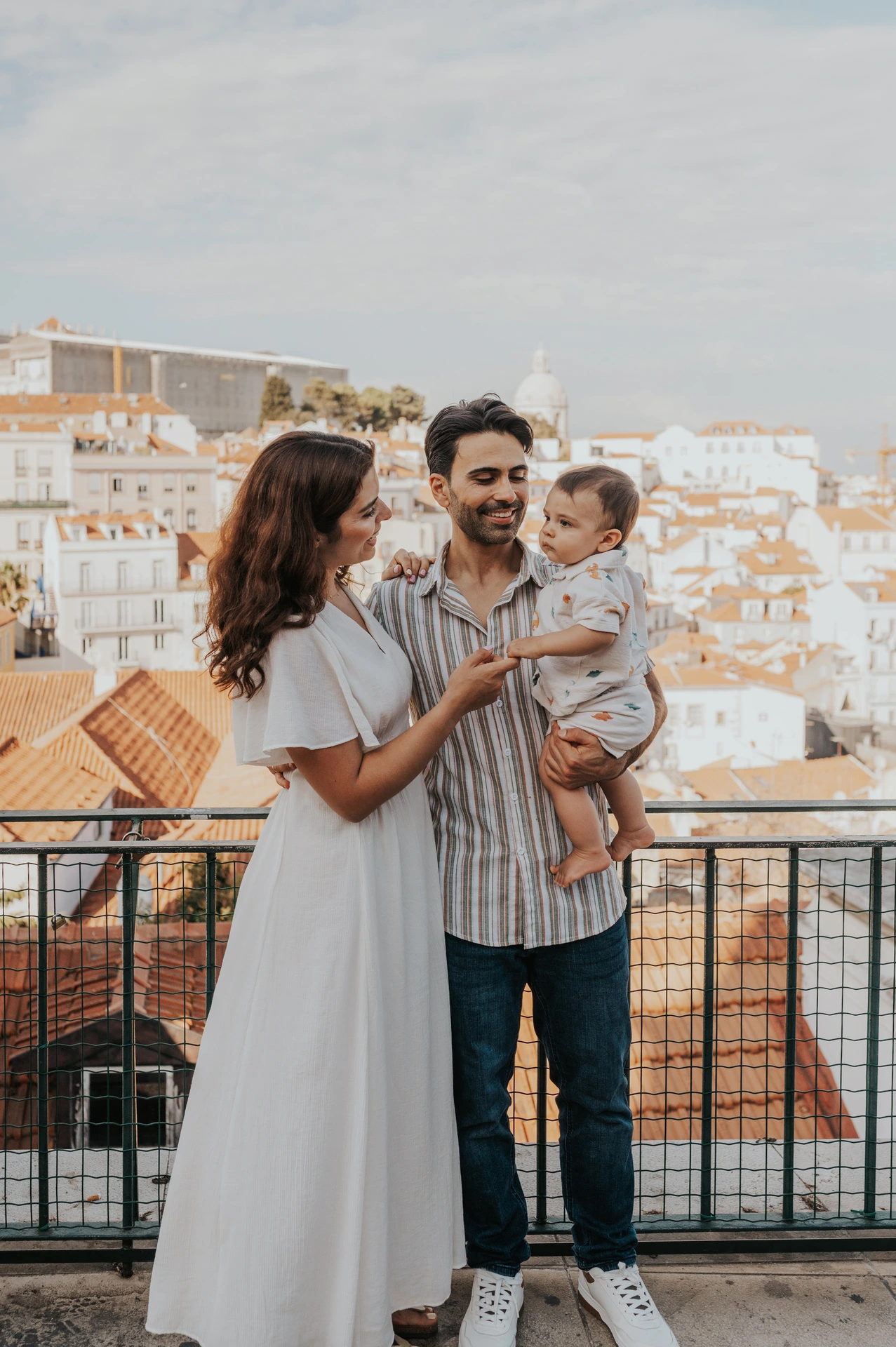 The parents holding their child together beside the viewpoint railing with Lisbon’s rooftops behind them during their family photoshoot in Lisbon.