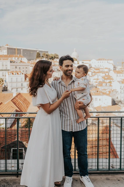 A family portrait above the rooftops | Lisbon family photoshoot The parents holding their child together beside the viewpoint railing with Lisbon’s rooftops behind them during their family photoshoot in Lisbon.
