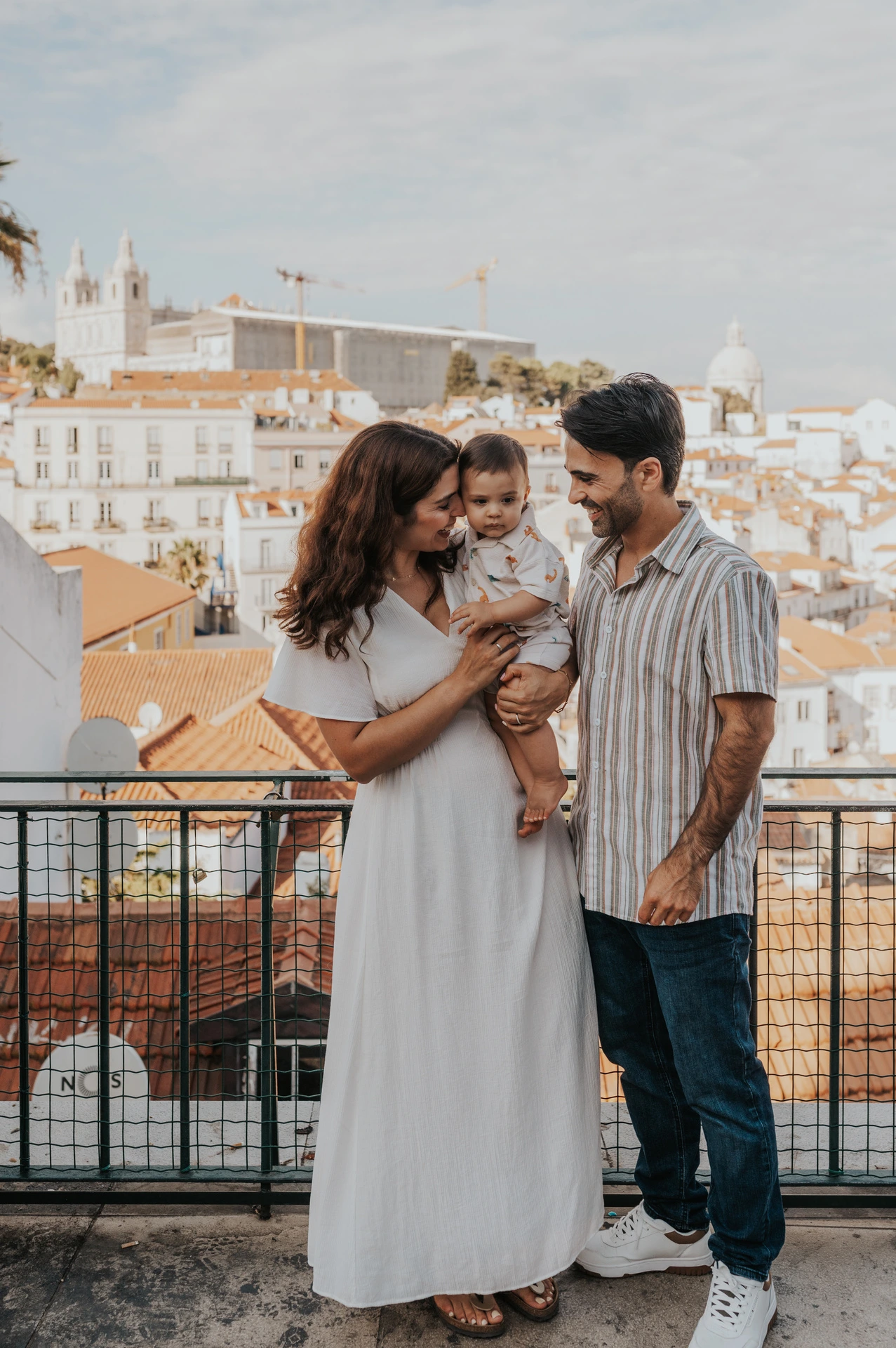 The parents holding their child together beside the viewpoint railing with Lisbon’s rooftops behind them during their family photoshoot in Lisbon.