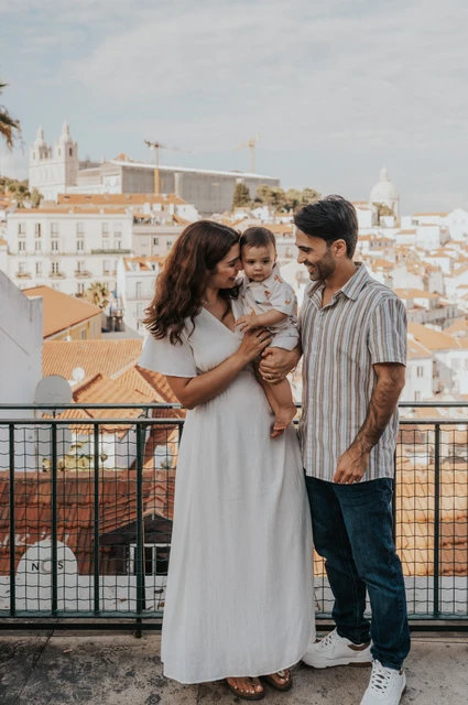 A family portrait above the rooftops | Lisbon family photoshoot The parents holding their child together beside the viewpoint railing with Lisbon’s rooftops behind them during their family photoshoot in Lisbon.