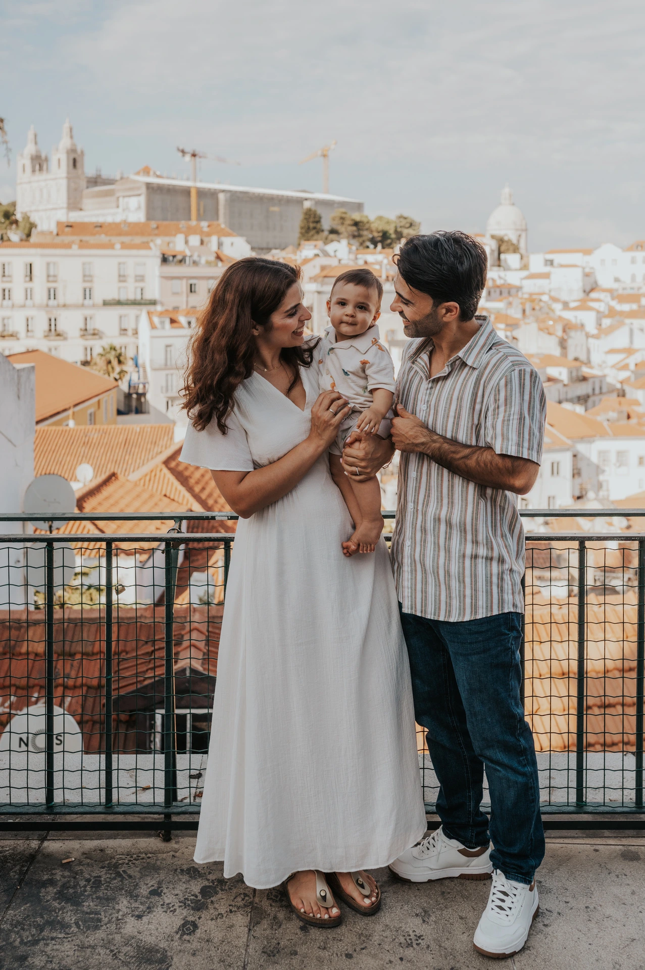 The parents holding their child together beside the viewpoint railing with Lisbon’s rooftops behind them during their family photoshoot in Lisbon.