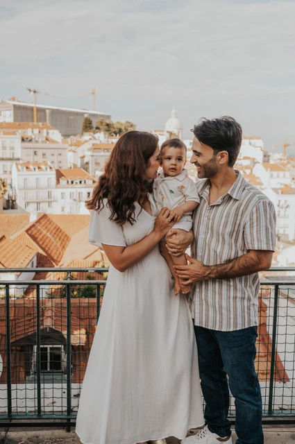 A family portrait above the rooftops | Lisbon family photoshoot The parents holding their child together beside the viewpoint railing with Lisbon’s rooftops behind them during their family photoshoot in Lisbon.