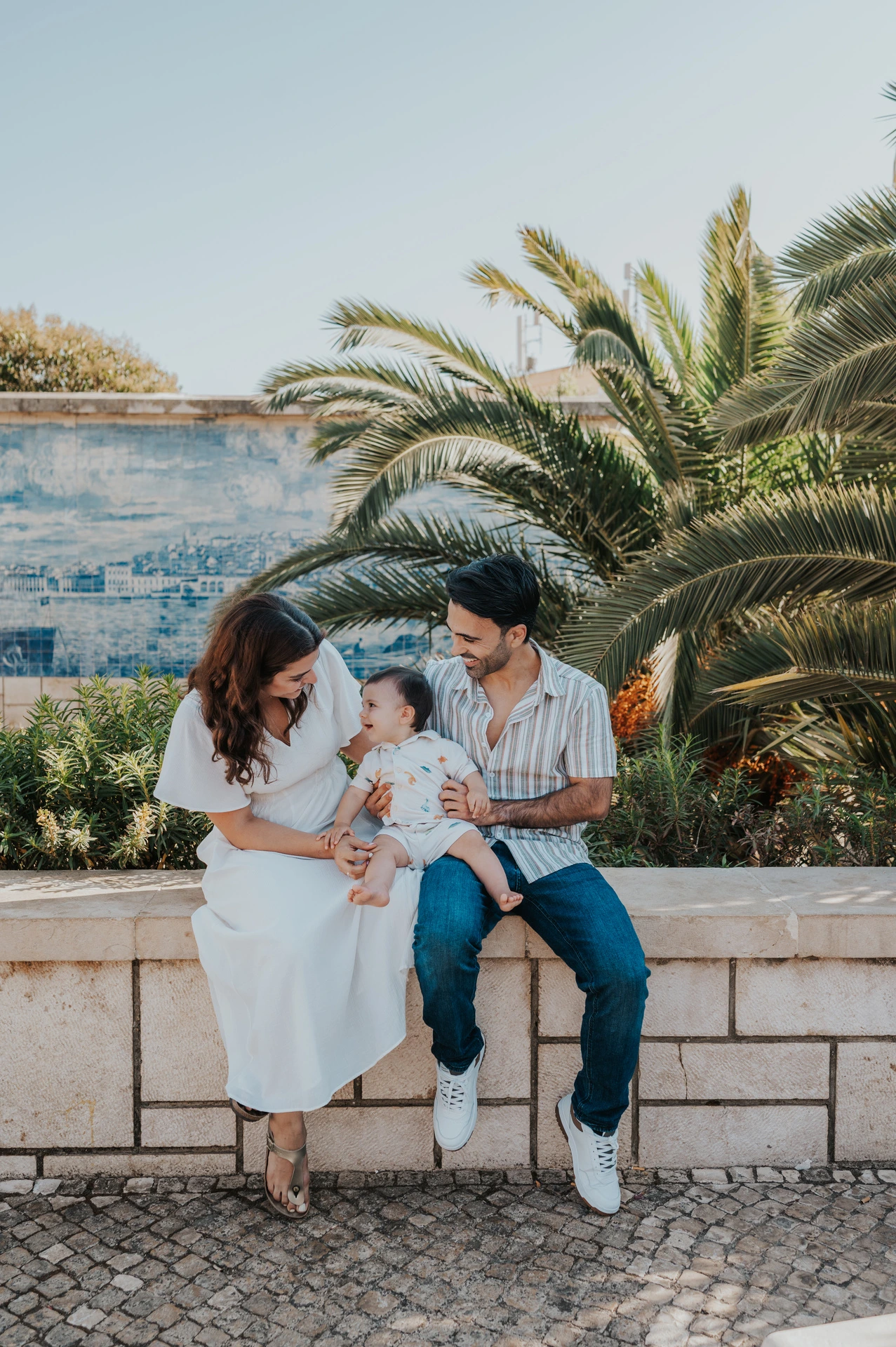 The parents seated together with their one-year-old child beside palms and blue Portuguese tiles during their family photoshoot in Lisbon.