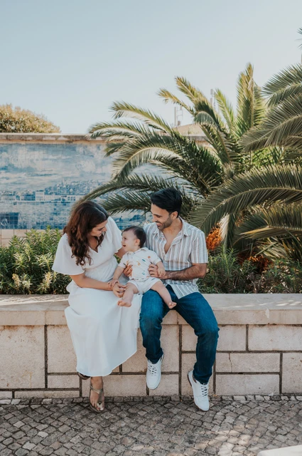 The parents seated together with their one-year-old child beside palms and blue Portuguese tiles during their family photoshoot in Lisbon.