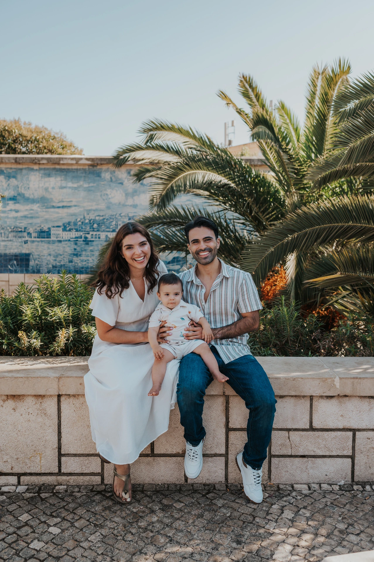 The parents seated together with their one-year-old child beside palms and blue Portuguese tiles during their family photoshoot in Lisbon.