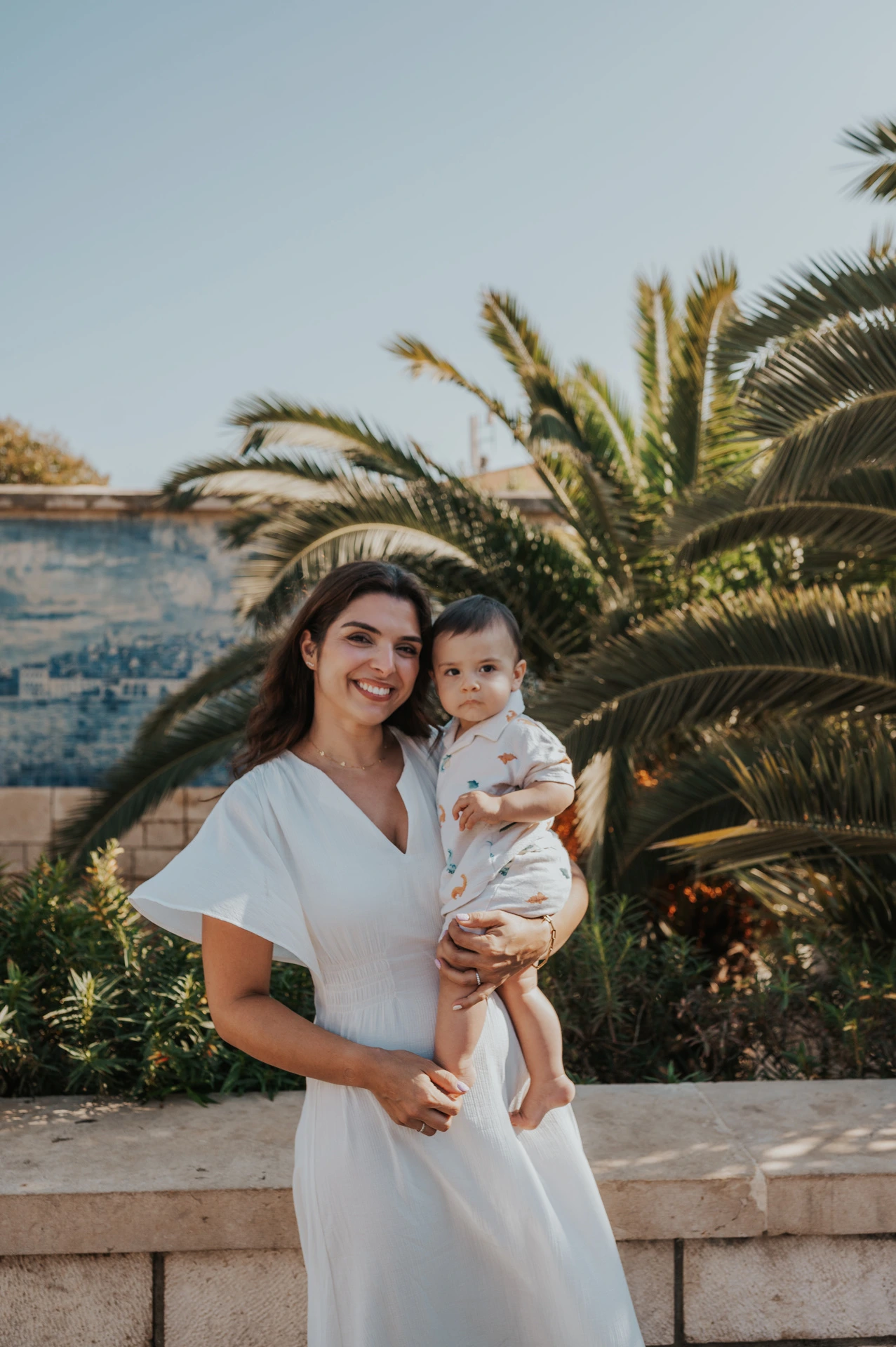 The mother holding her child close beside the palms and blue tile wall during their family photoshoot in Lisbon.