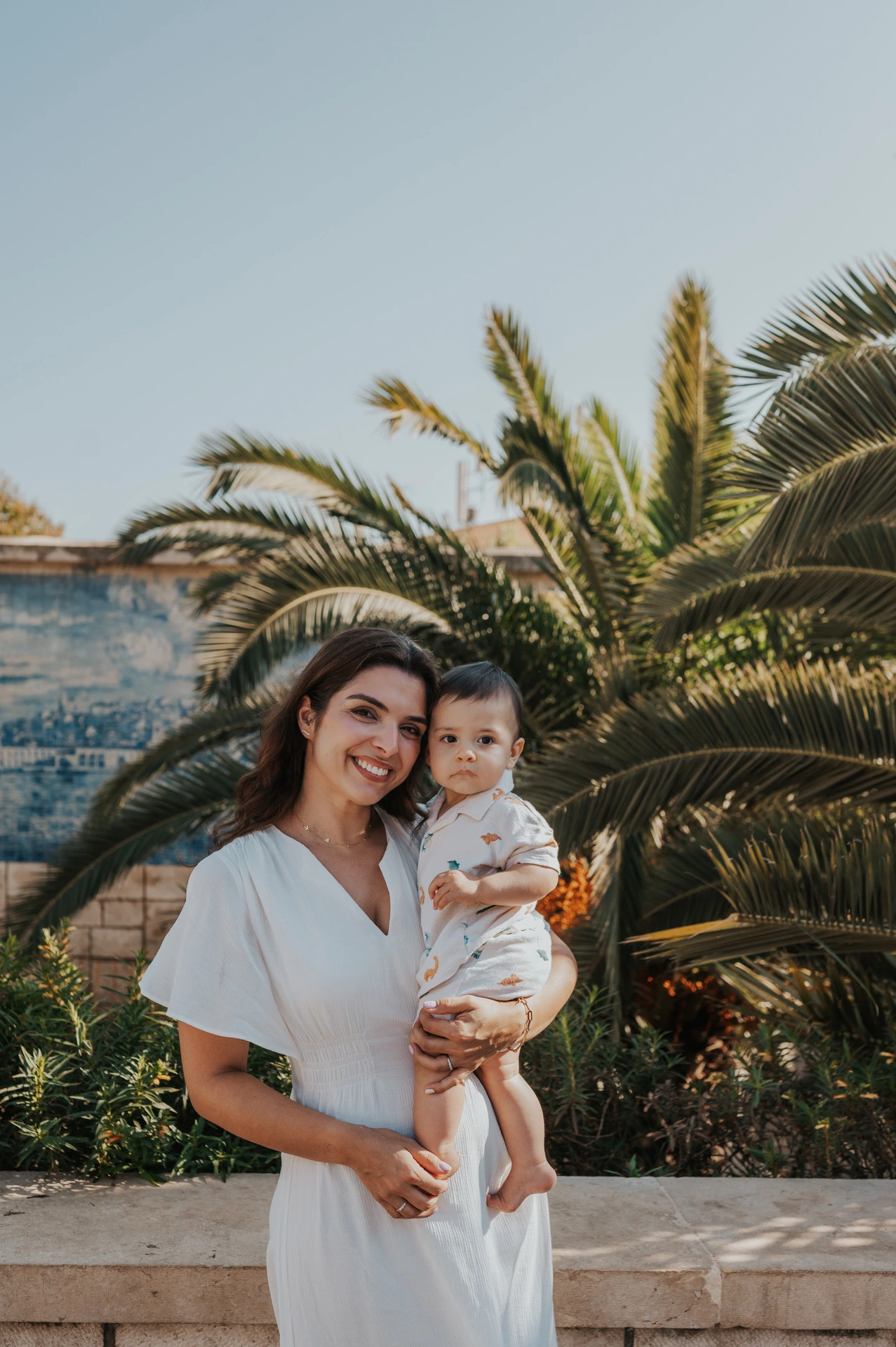 The mother holding her child close beside the palms and blue tile wall during their family photoshoot in Lisbon.