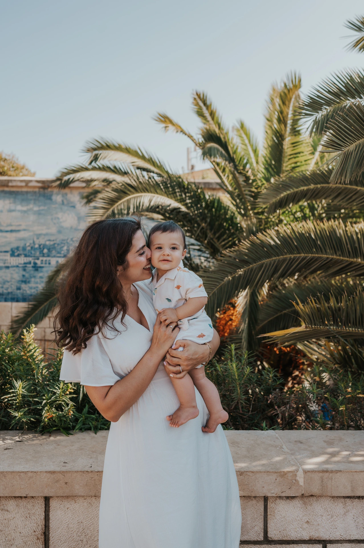 The mother holding her child close beside the palms and blue tile wall during their family photoshoot in Lisbon.