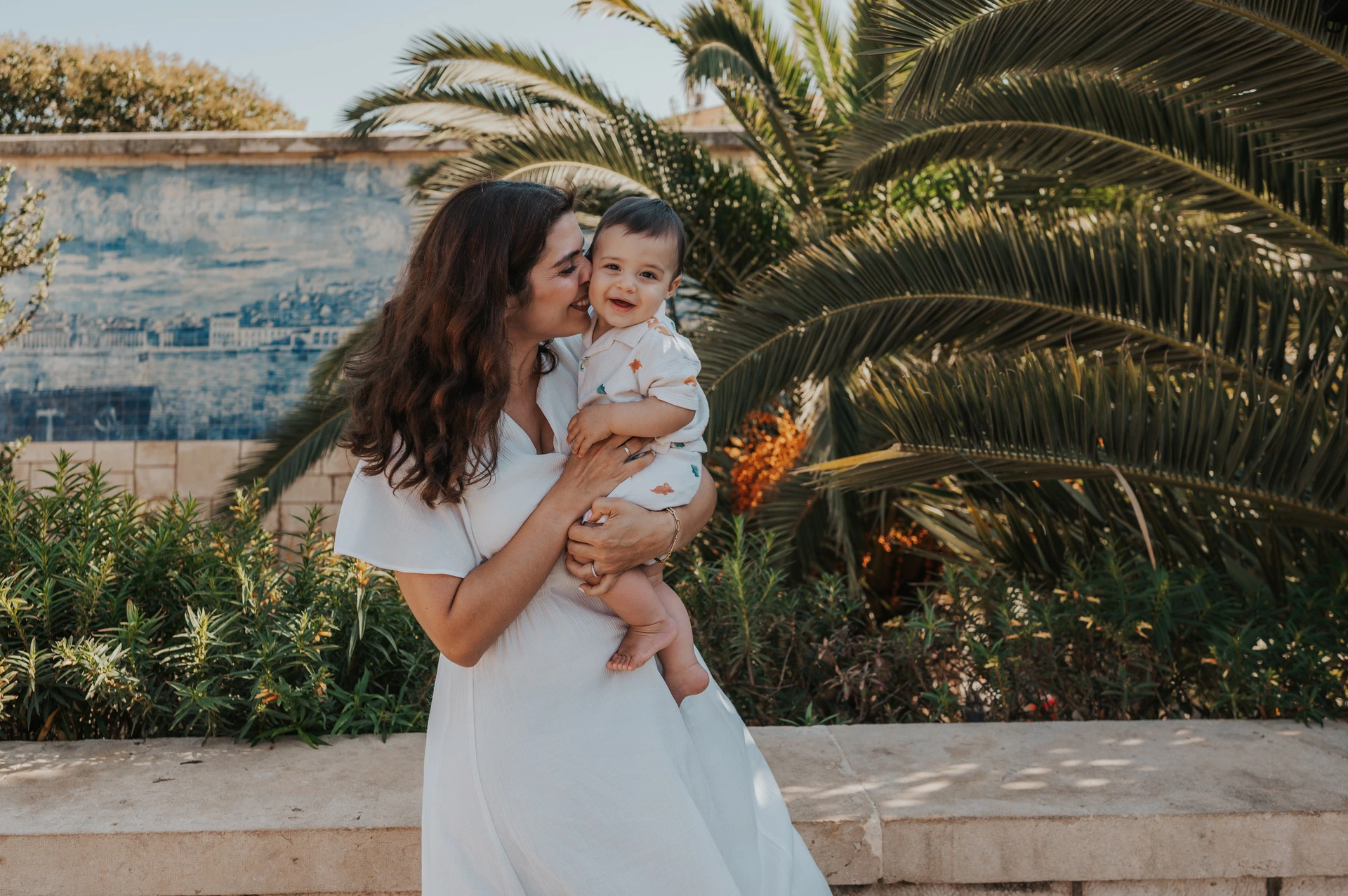 The mother holding her child close beside the palms and blue tile wall during their family photoshoot in Lisbon.