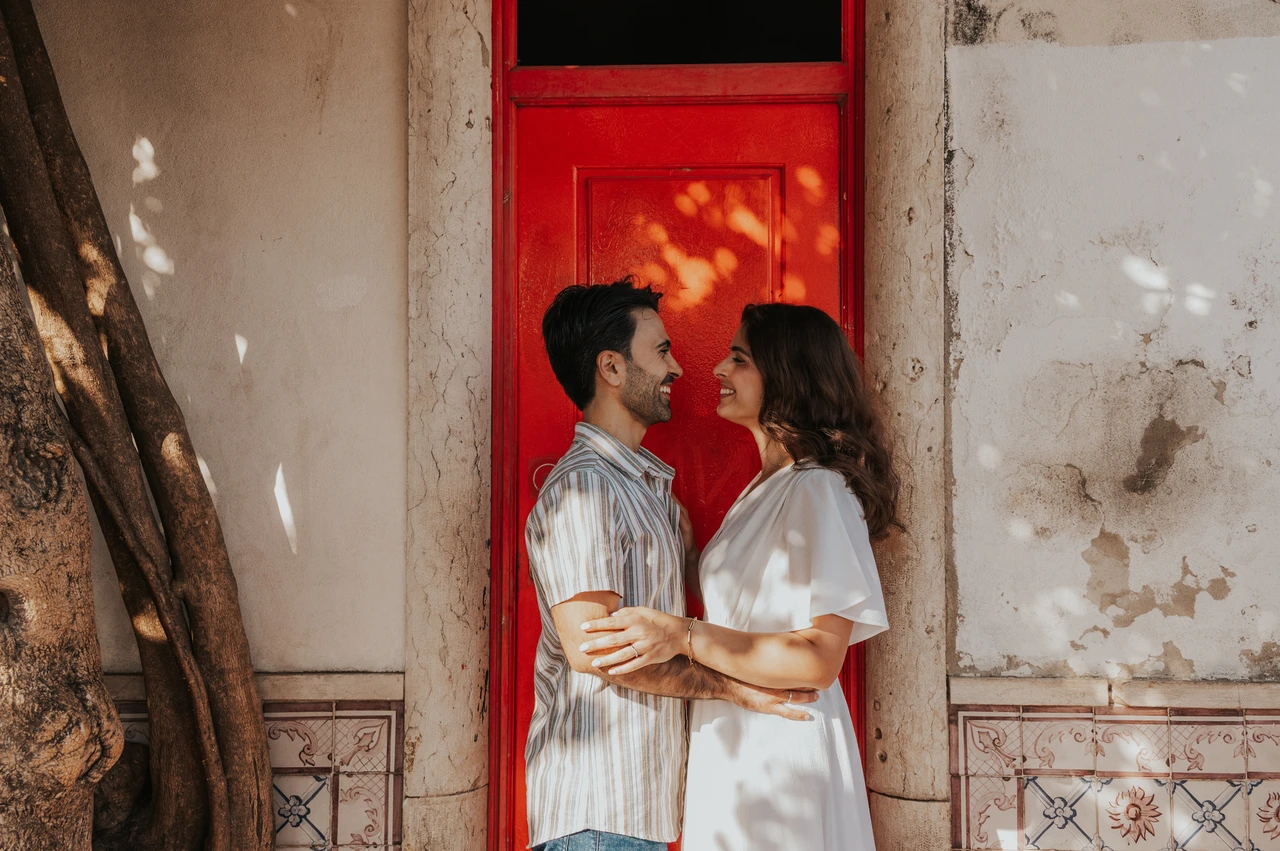 A close portrait of the parents at the red door | Lisbon family photoshoot The parents standing close together and facing each other beside the red doorway during their family photoshoot in Lisbon.