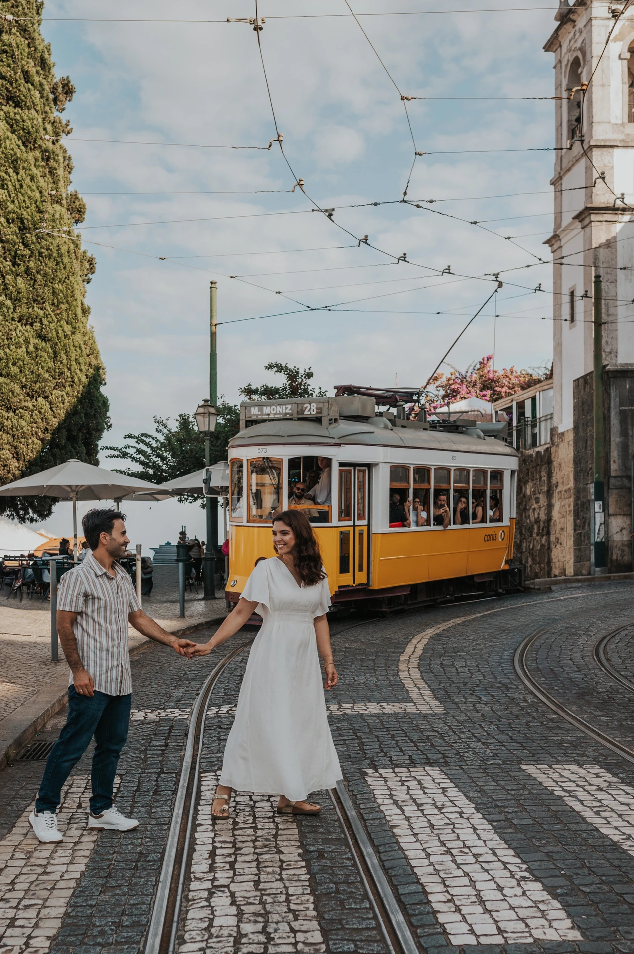The parents holding hands beside a Lisbon street with a yellow tram behind them during their family photoshoot in Lisbon.