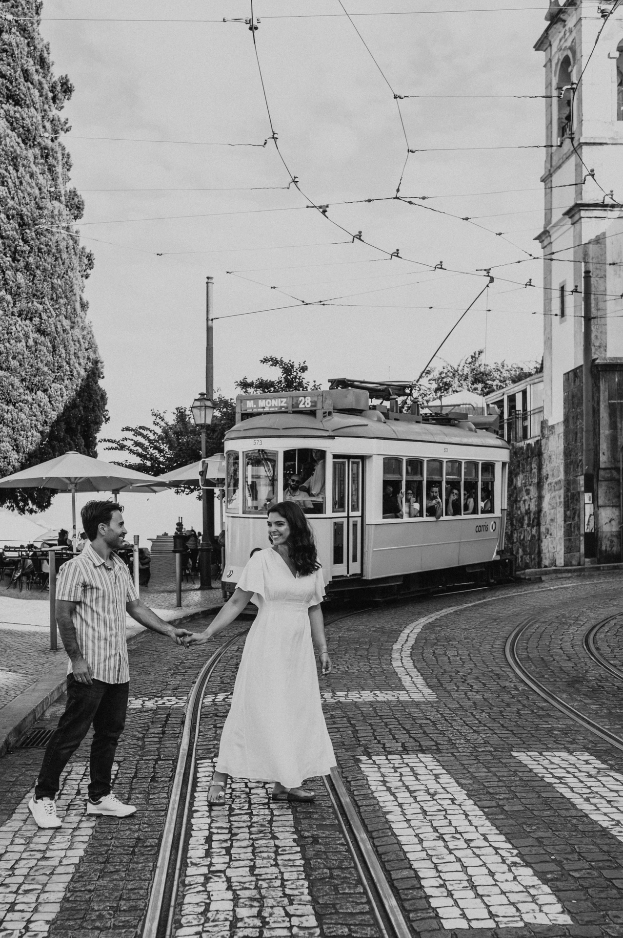 Black-and-white photograph: the parents holding hands beside a Lisbon street with a yellow tram behind them during their family photoshoot in Lisbon.
