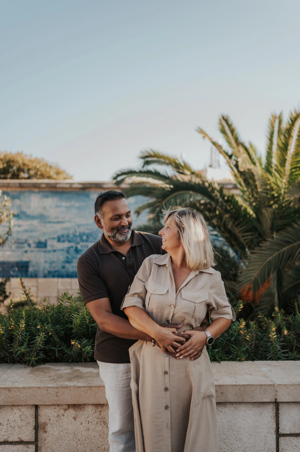 A smiling embrace beside the palms | Lisbon couple photoshoot The man embracing his smiling partner from behind beside palms and blue tiles during their couple photoshoot in Lisbon.