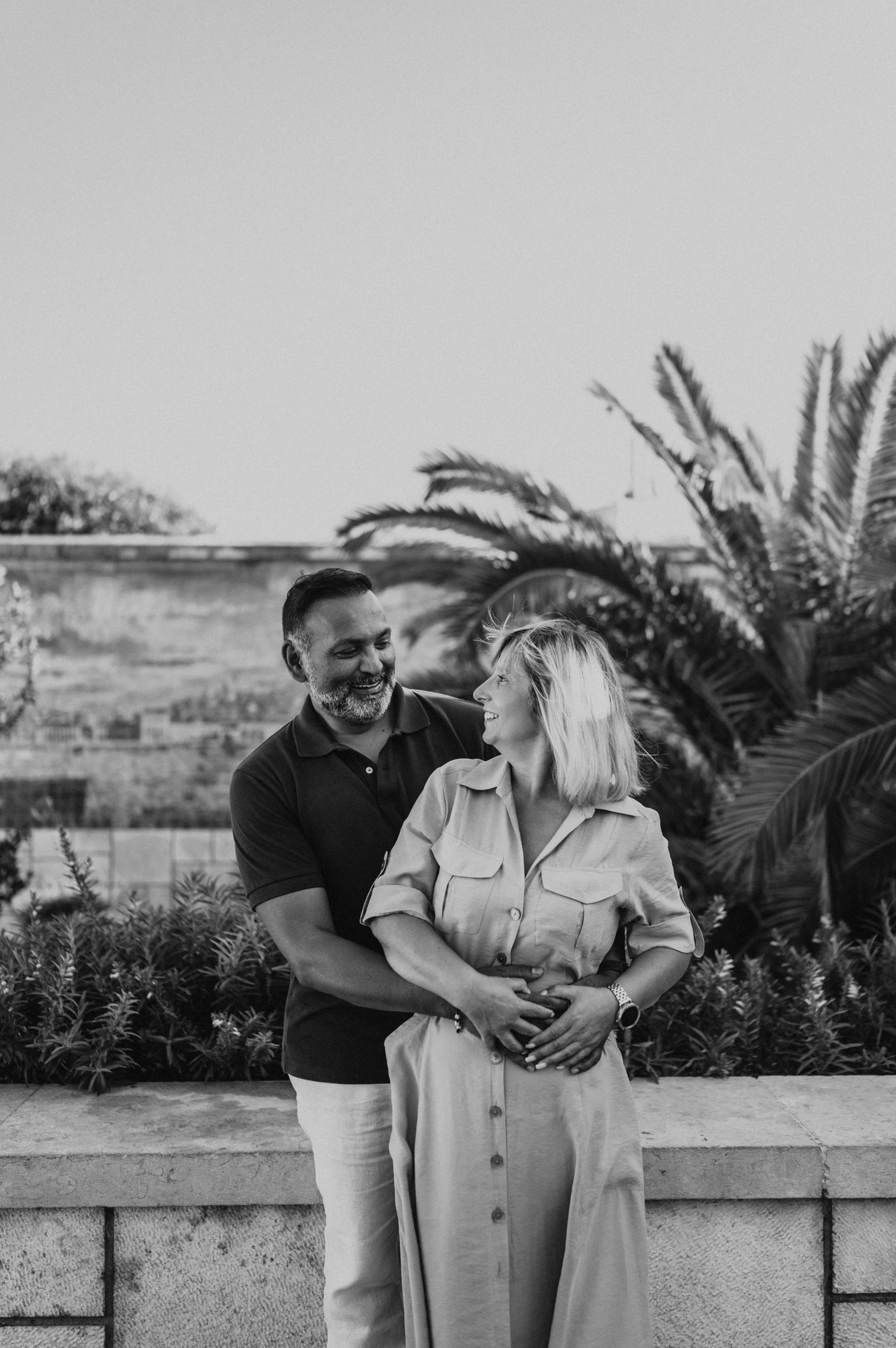 Black-and-white photograph: the man embracing his smiling partner from behind beside palms and blue tiles during their couple photoshoot in Lisbon.