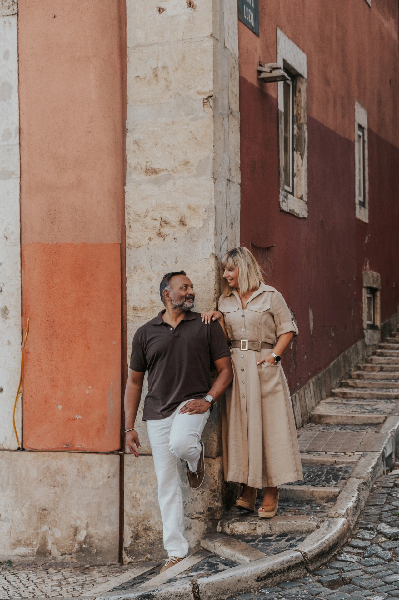 The couple holding hands beside the corner of a red building on a cobbled street during their couple photoshoot in Lisbon.