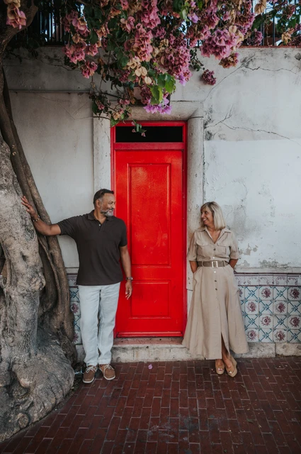 The couple standing apart beside a bright red door beneath flowering branches during their couple photoshoot in Lisbon.