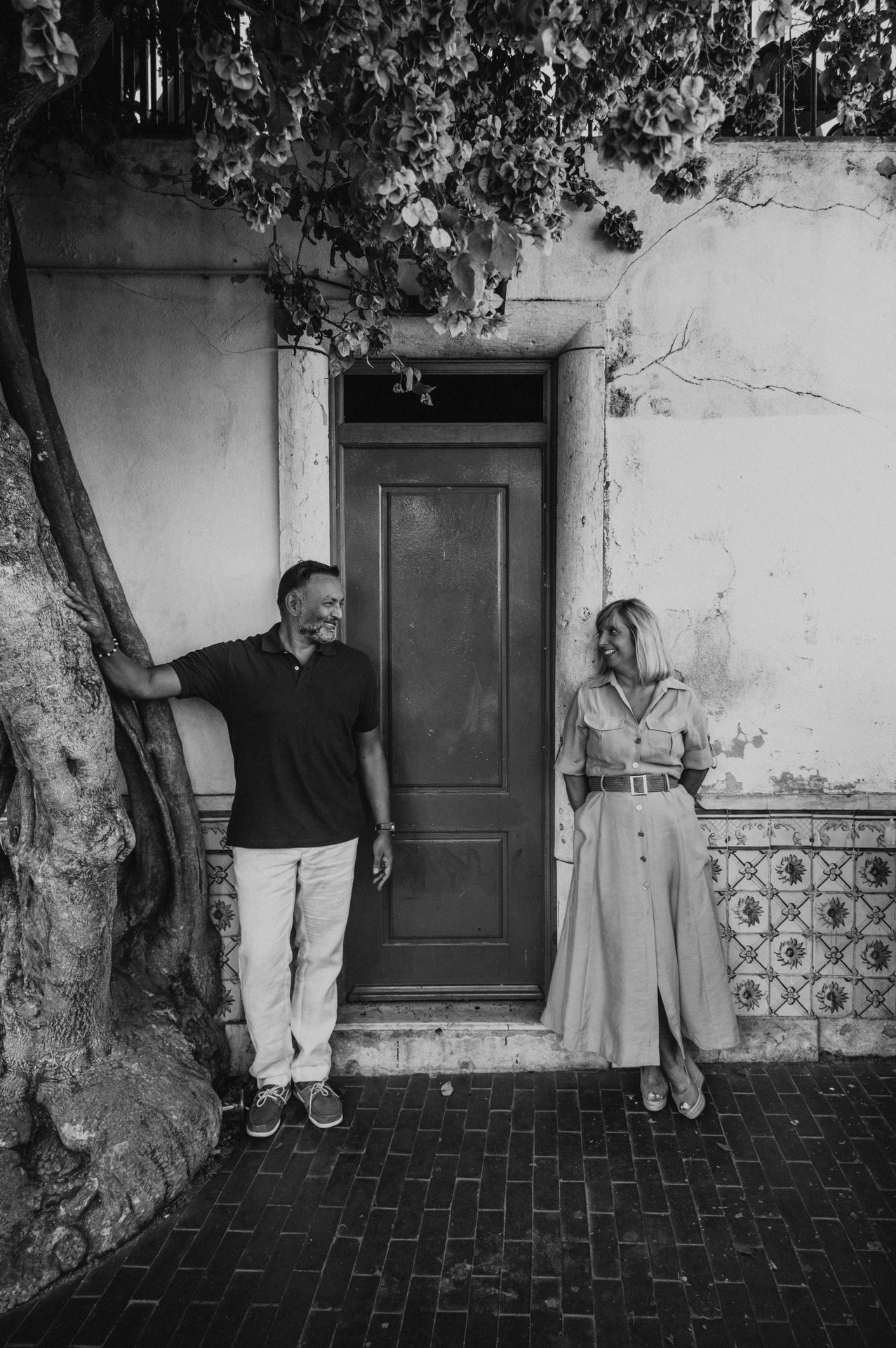 Black-and-white photograph: the couple standing apart beside a bright red door beneath flowering branches during their couple photoshoot in Lisbon.