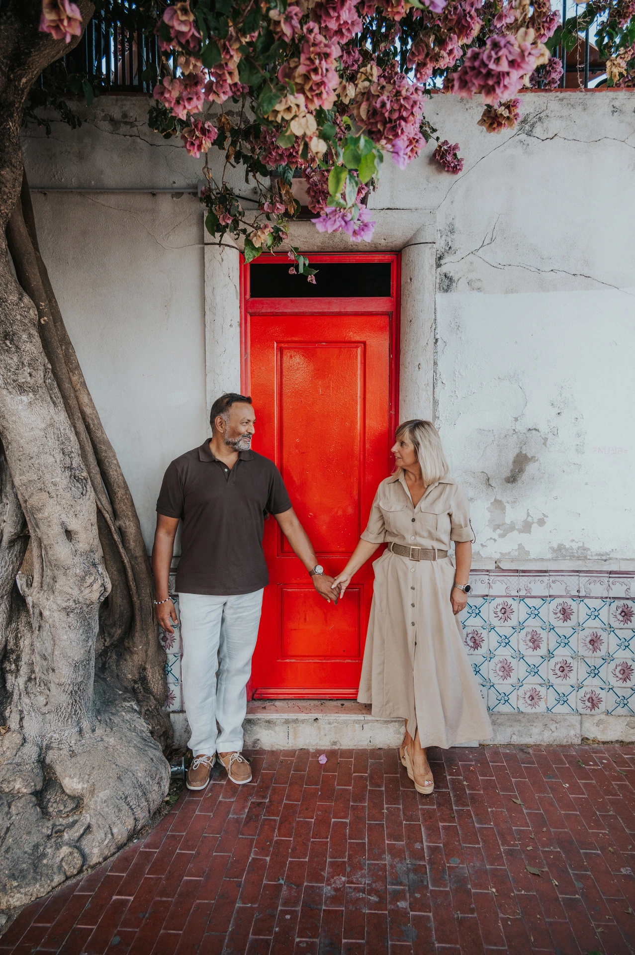 The couple holding hands across the red doorway beneath flowering branches during their couple photoshoot in Lisbon.