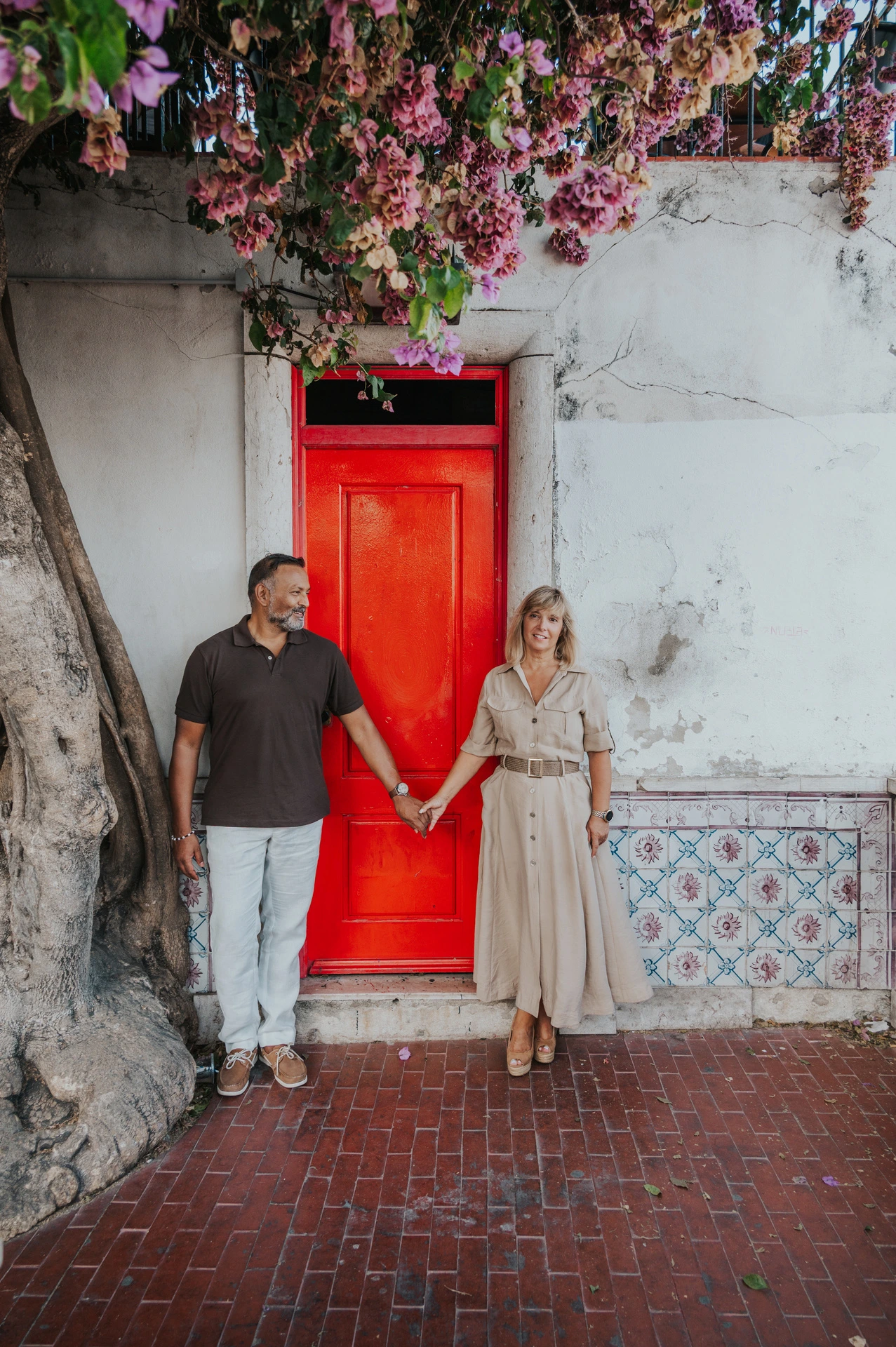 The couple holding hands across the red doorway beneath flowering branches during their couple photoshoot in Lisbon.