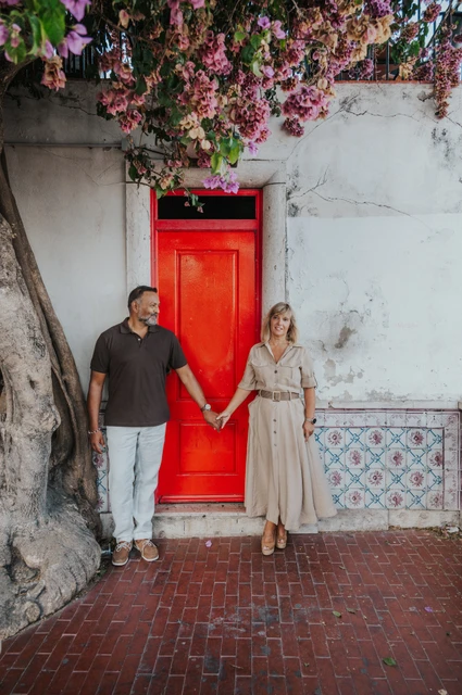 Joined hands beside the red door | Lisbon couple photoshoot The couple holding hands across the red doorway beneath flowering branches during their couple photoshoot in Lisbon.