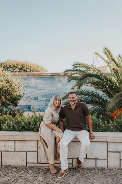Seated together beside the palms and tiles | Lisbon couple photoshoot The couple seated close together on a low wall beside palm leaves and blue Portuguese tiles during their couple photoshoot in Lisbon.