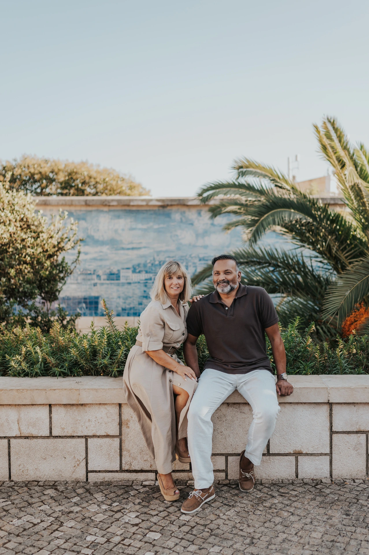 The couple seated close together on a low wall beside palm leaves and blue Portuguese tiles during their couple photoshoot in Lisbon.