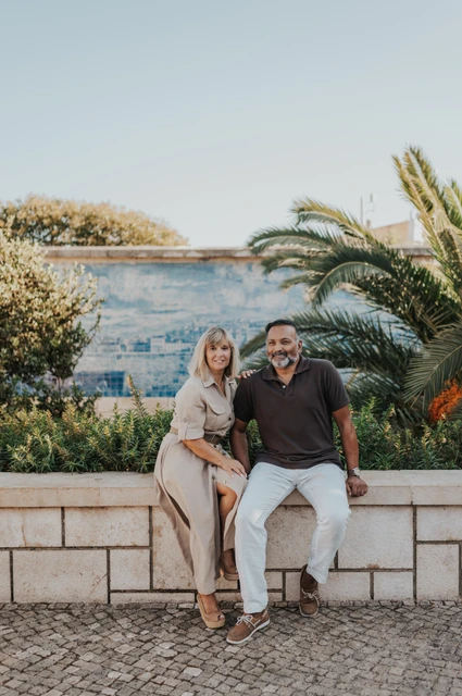 Seated together beside the palms and tiles | Lisbon couple photoshoot The couple seated close together on a low wall beside palm leaves and blue Portuguese tiles during their couple photoshoot in Lisbon.