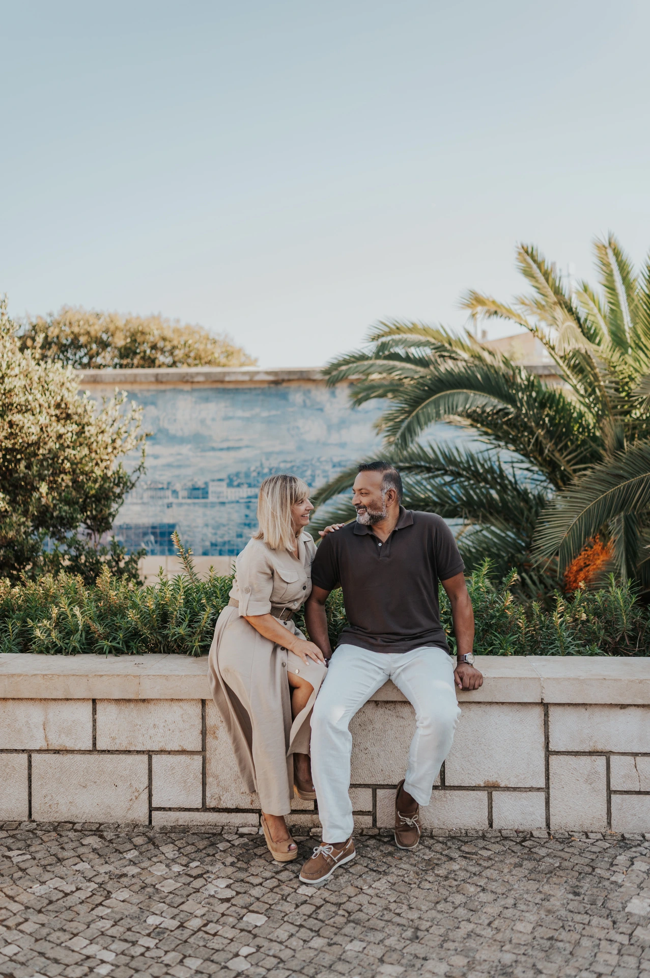The couple seated close together on a low wall beside palm leaves and blue Portuguese tiles during their couple photoshoot in Lisbon.