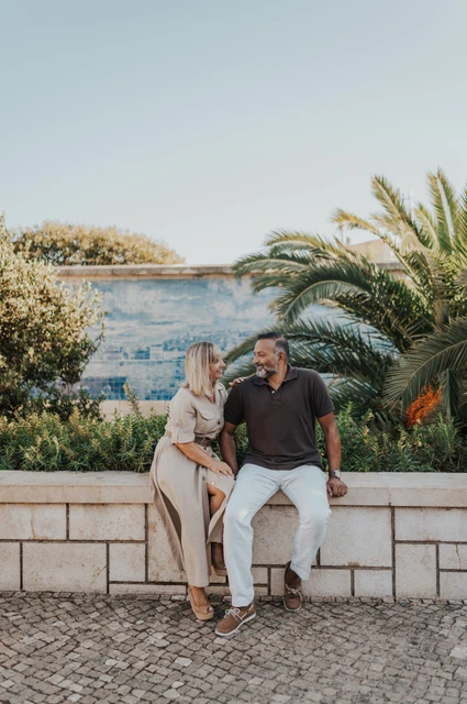Seated together beside the palms and tiles | Lisbon couple photoshoot The couple seated close together on a low wall beside palm leaves and blue Portuguese tiles during their couple photoshoot in Lisbon.
