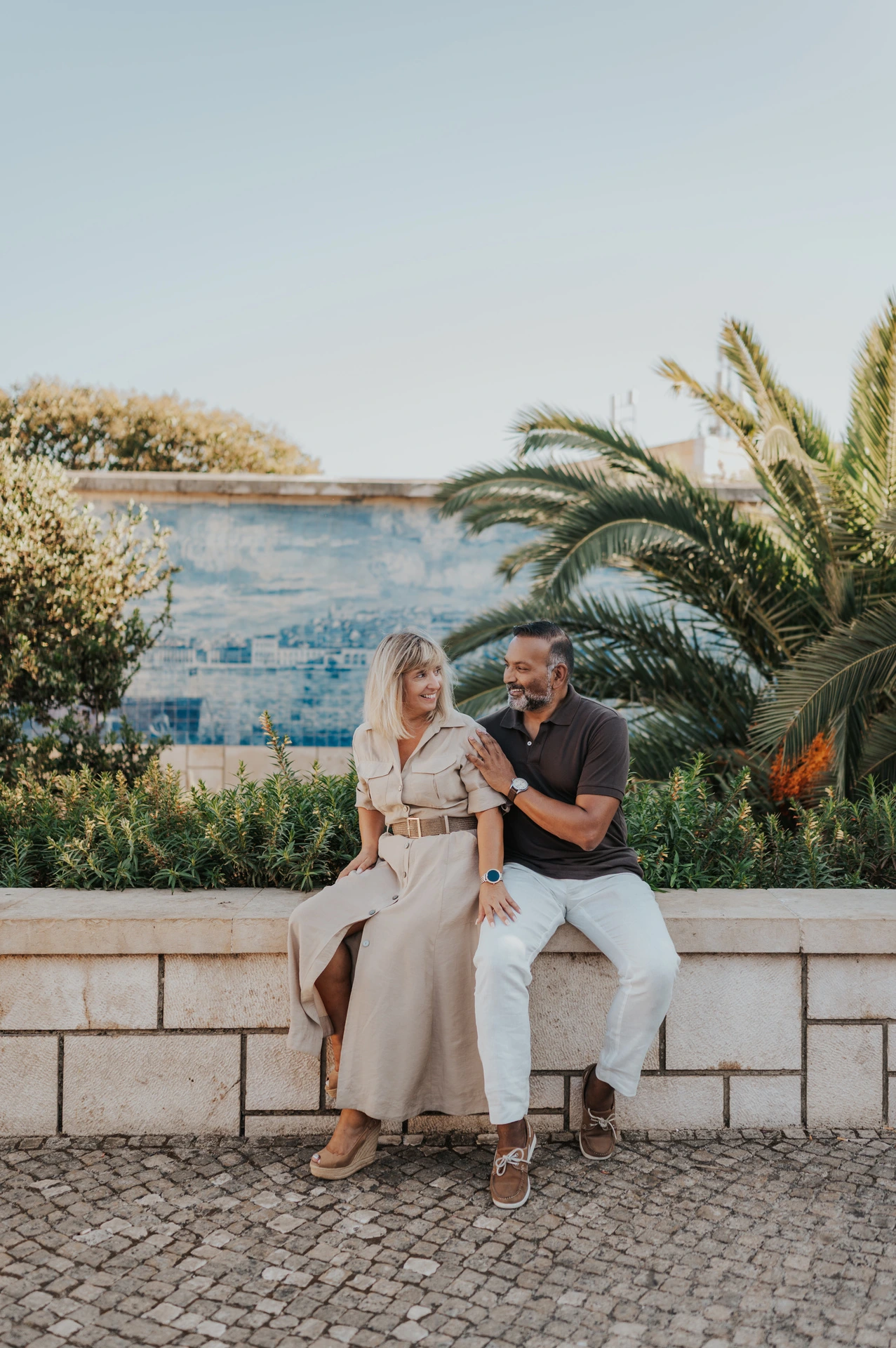 The couple seated close together on a low wall beside palm leaves and blue Portuguese tiles during their couple photoshoot in Lisbon.