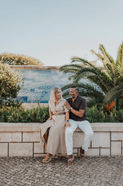 Seated together beside the palms and tiles | Lisbon couple photoshoot The couple seated close together on a low wall beside palm leaves and blue Portuguese tiles during their couple photoshoot in Lisbon.