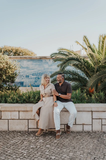 Seated together beside the palms and tiles | Lisbon couple photoshoot The couple seated close together on a low wall beside palm leaves and blue Portuguese tiles during their couple photoshoot in Lisbon.
