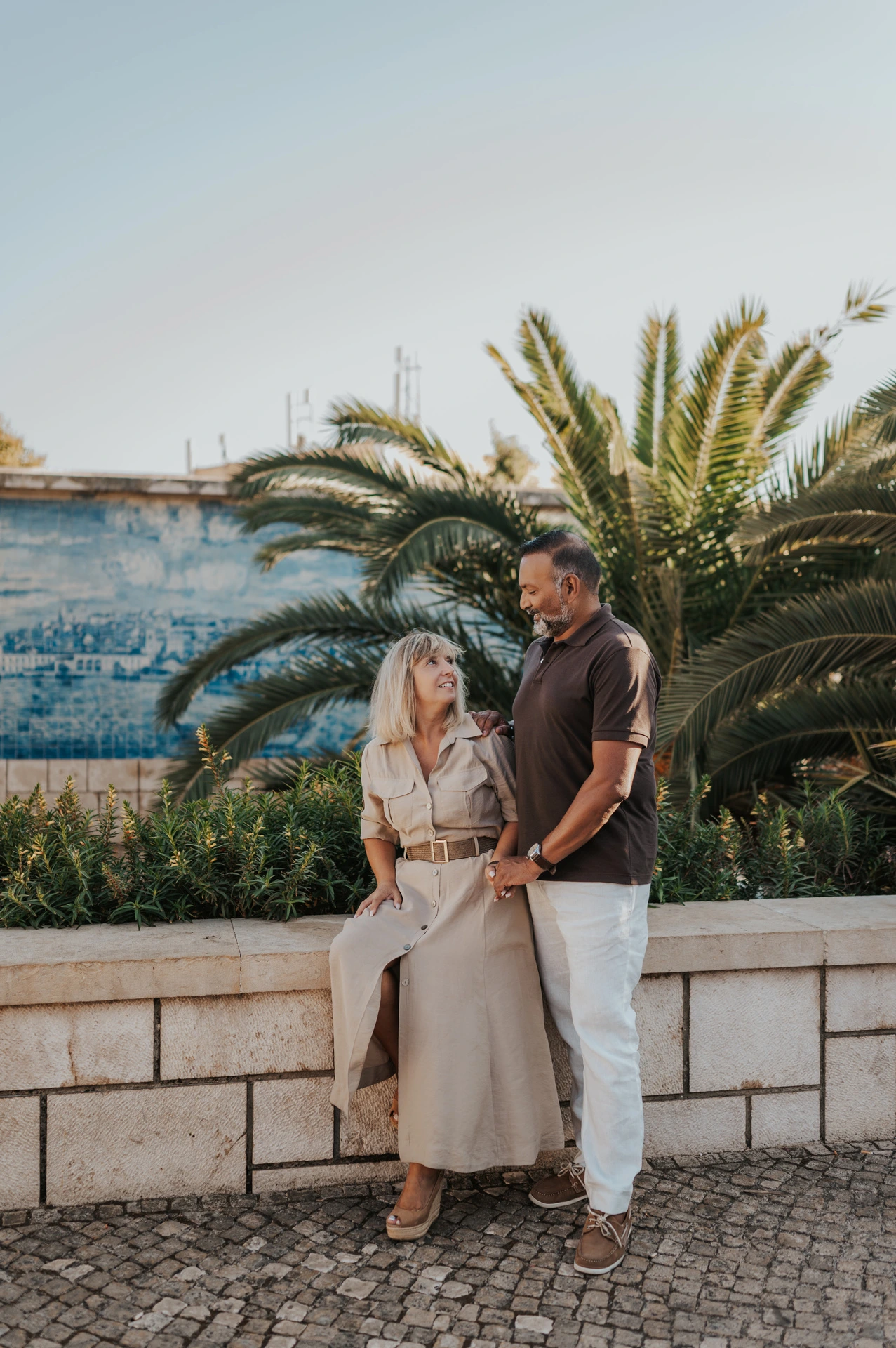 The woman seated on the low wall with her partner standing close beside her by the tiled viewpoint during their couple photoshoot in Lisbon.