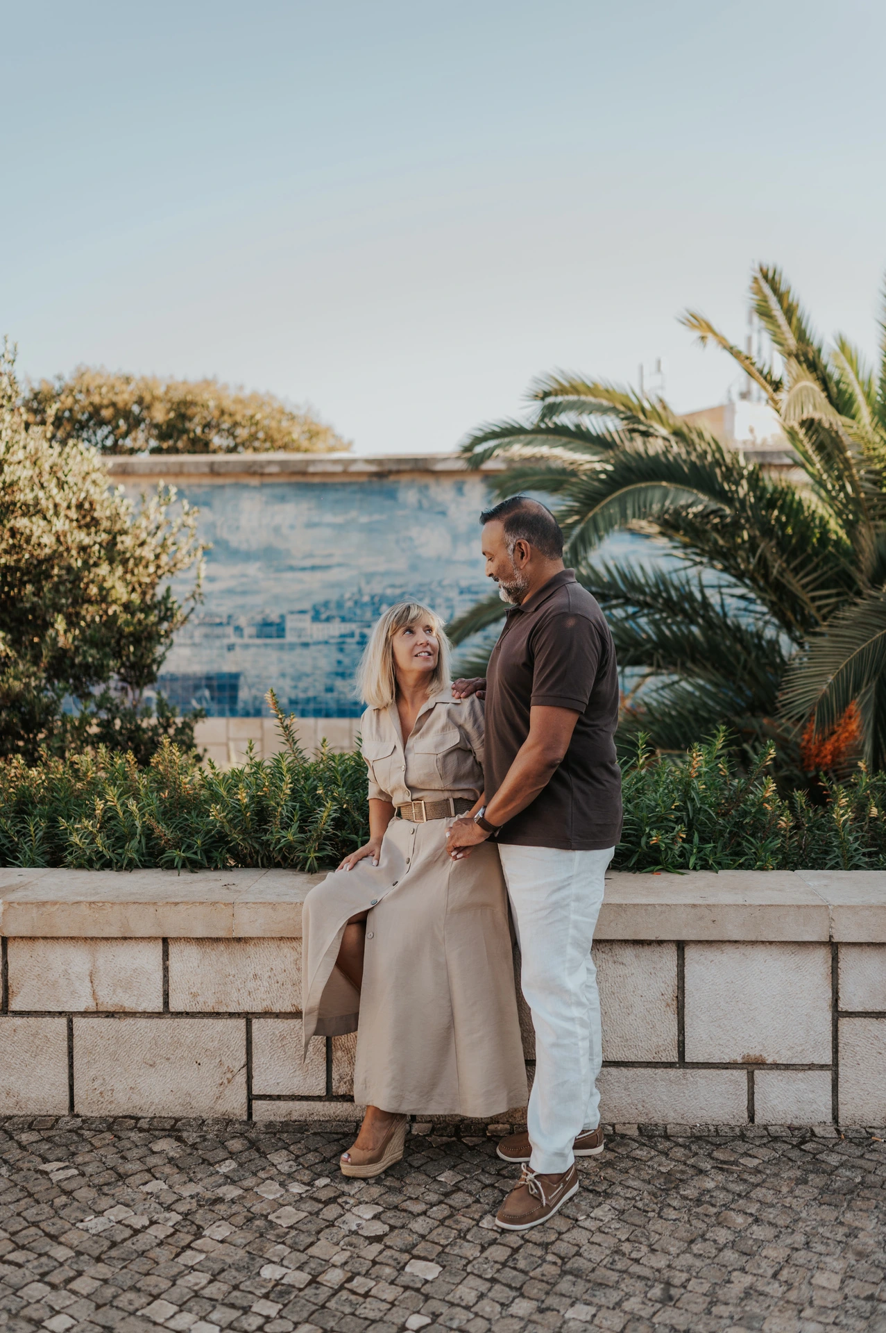 The woman seated on the low wall with her partner standing close beside her by the tiled viewpoint during their couple photoshoot in Lisbon.