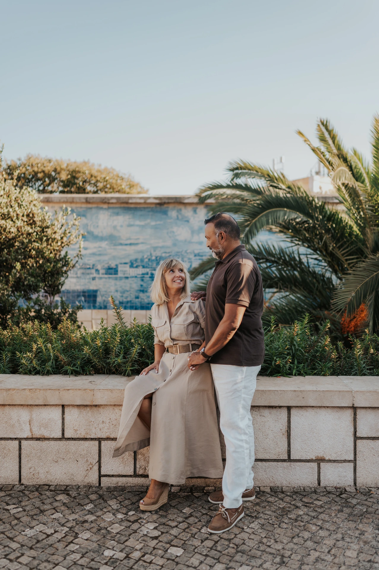 The woman seated on the low wall with her partner standing close beside her by the tiled viewpoint during their couple photoshoot in Lisbon.