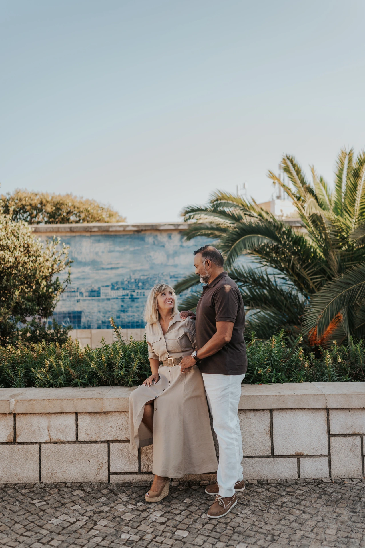 The woman seated on the low wall with her partner standing close beside her by the tiled viewpoint during their couple photoshoot in Lisbon.