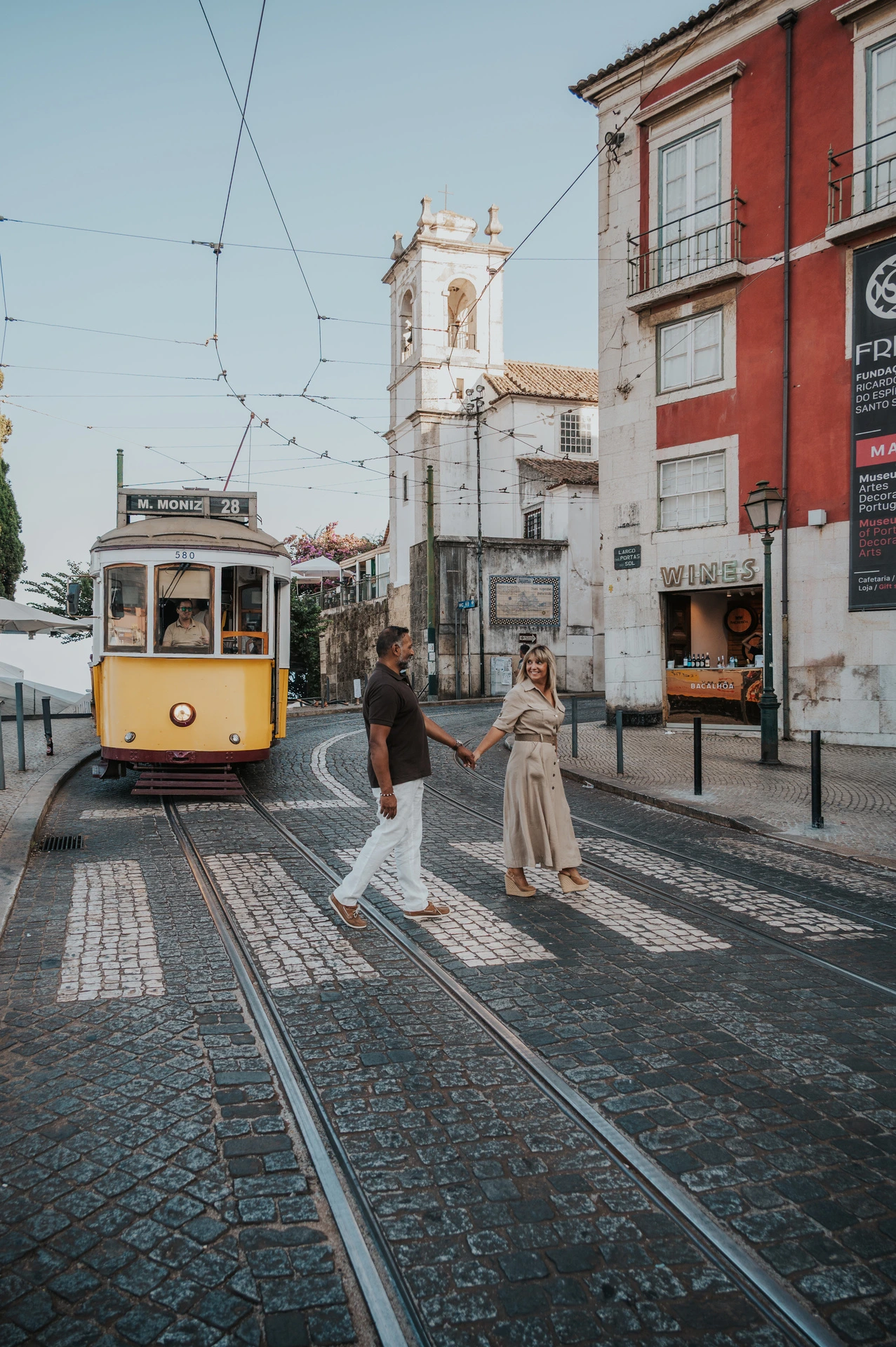The couple standing together beside a Lisbon street with a yellow tram behind them during their couple photoshoot in Lisbon.